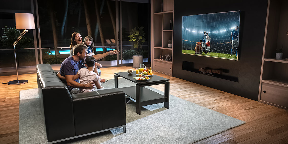 A man, woman and two children watch football on a large tv in a spacious living room