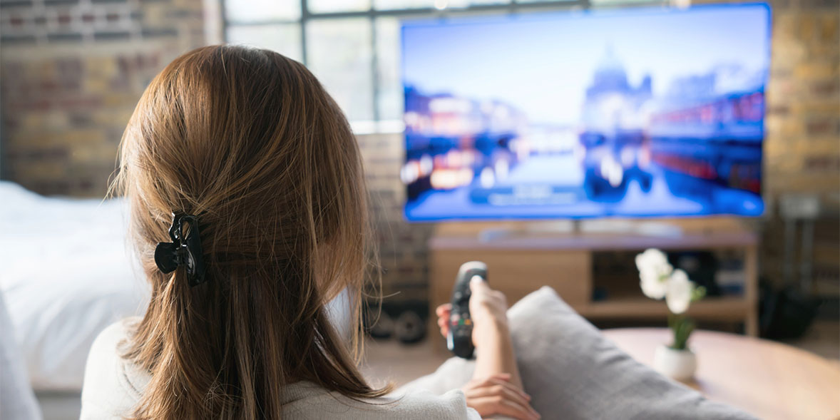 Woman sitting on a sofa, remote in watching a silver television on a wooden media unit