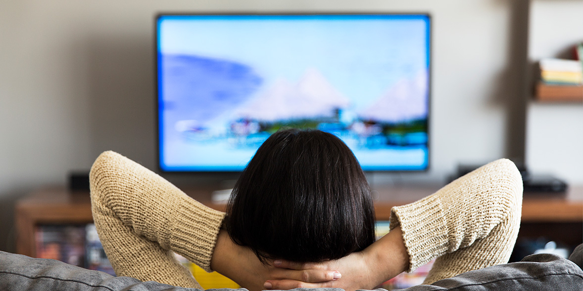 A woman on her sofa watching TV with her arms behind her head in a relaxed pose