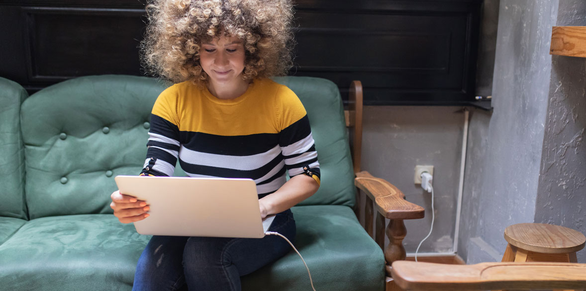A person sitting on a sofa using a laptop that is plugged into the wall