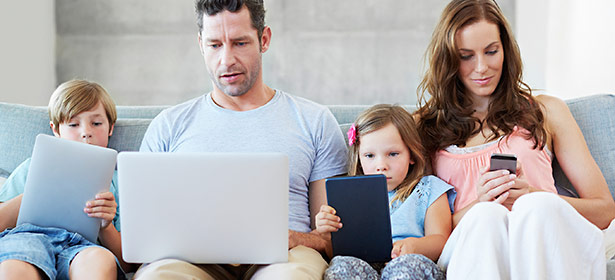 A family - two parents and two children - sitting on their sofa while all using different connected devices.